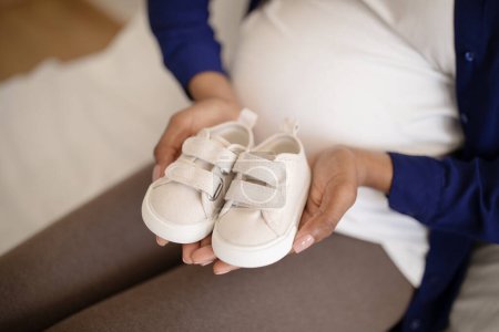 Photo for Millennial african american lady with big belly hold small shoes for future baby in room interior, cropped, close up. Childbirth, expectation of child, motherhood and family - Royalty Free Image