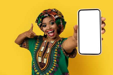 Téléchargez les photos : Belle nouvelle application mobile. Émotionnel attrayant jeune femme noire dans un habillage africain coloré lumineux montrant smartphone moderne avec écran blanc vierge, maquette, fond jaune - en image libre de droit