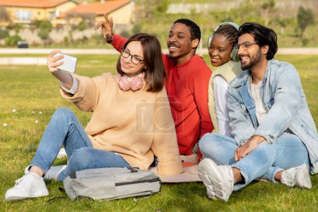 Téléchargez les photos : Heureux étudiants millénaires multiethniques étudient, s'asseoir sur l'herbe, prendre selfie ensemble pour blog à l'université en plein air. Photo pour les réseaux sociaux, pause et éducation, connaissance et communication - en image libre de droit