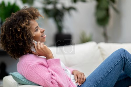 Téléchargez les photos : Relaxé joyeux jolie jeune femme noire bouclée portant tenue décontractée allongé sur le canapé à la maison, parlant au téléphone et souriant, regardant l'espace de copie, concept de communication - en image libre de droit