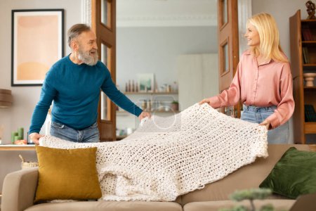 Photo for Timeless Comfort. Joyful Senior Spouses Covering Couch With New Knitted Blanket, Adding A Touch Of Love To Their Home. Couple Tidying Living Room Doing House Chores Together. Housekeeping Leisure - Royalty Free Image