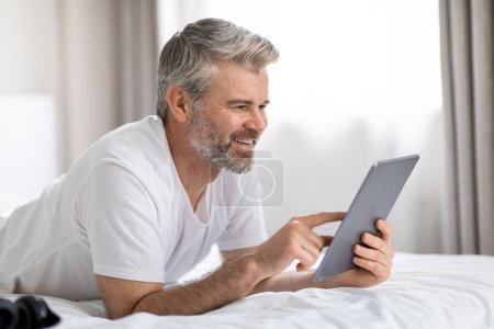 Photo for Joyful attractive grey-haired bearded middle aged man in white t-shirt chilling in bed after waking up in the morning, using digital tablet and smiling, scrolling on social media, copy space - Royalty Free Image