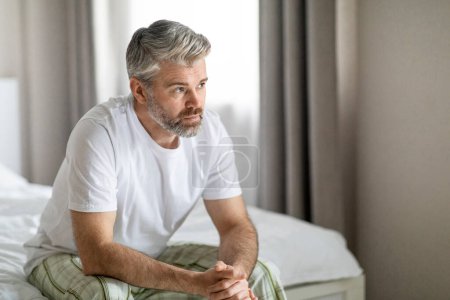 Photo for Pensive thoughtful unhappy handsome grey-haired bearded mature caucasian man wearing pajamas sitting on bed at home, looking at copy space, feeling lonely, going through divorce - Royalty Free Image