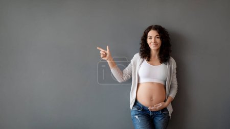 Photo for Smiling expectant mother pointing to copy space beside her and tenderly touching belly, young pregnant female standing near grey wall, demonstrating background for your advertisement, panorama - Royalty Free Image