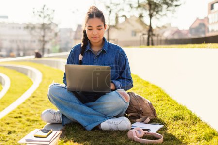 Female mixed race student sitting with laptop in urban park, learning online, having educational virtual lecture outdoors. Modern study and education concept