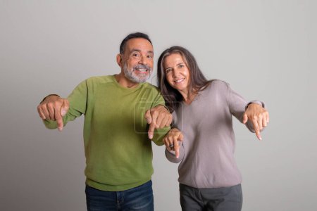 Photo for Cheerful caucasian senior man and lady pointing fingers down at copy space recommending ad and offer on gray studio background. Sale, advice and fun together, people emotions - Royalty Free Image