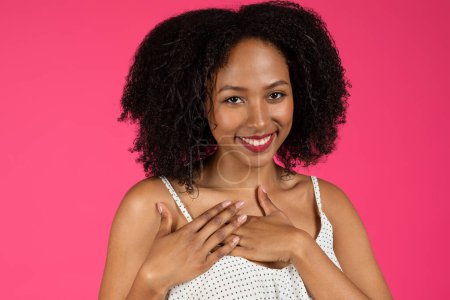 Foto de Retrato de alegre linda mujer rizada afroamericana joven poner las manos en el pecho, hacer signo de agradecimiento, aislado en el fondo del estudio rosa, de cerca. Gracias gesto, amor, anuncio y oferta - Imagen libre de derechos