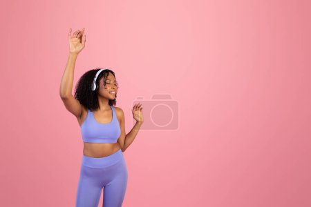 Photo for Glad millennial black woman in sportswear, wireless headphones with closed eyes enjoy music, dancing isolated on pink background, studio. Ad and offer, audio app for sports, fitness, pilates - Royalty Free Image