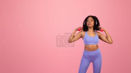 Photo for Serious millennial african american lady in sportswear and headphones do exercises with hand dumbbells isolated on pink background, studio. Sports with music, ad and offer, fitness and body care - Royalty Free Image