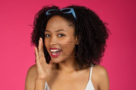 Photo for Happy cute young african american curly lady in sunglasses puts her hand to mouth and whispers secret, isolated on pink studio background, close up. Gossip gesture, communication, sale, good news - Royalty Free Image