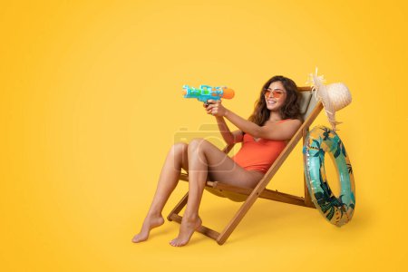 Foto de Encantada dama asiática milenaria en traje de baño, gafas de sol se sientan en la silla de cubierta, disfrutando del tiempo libre y jugando pistola de agua aislada en el fondo del estudio naranja. Diversión de verano, descanso y relax, tiempo libre - Imagen libre de derechos