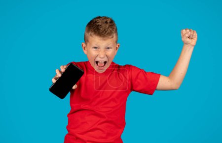 Photo for Overjoyed Little Boy Holding Smartphone With Blank Screen And Celebrating Success, Emotional Preteen Male Kid Demonstrating Empty Cellphone And Yelling With Excitement, Blue Background, Mockup - Royalty Free Image
