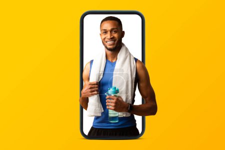 Photo for Positive millennial african american muscular man in sportswear with towel and water bottle on phone screen isolated on yellow studio background. Sport blog, body care app, workout - Royalty Free Image