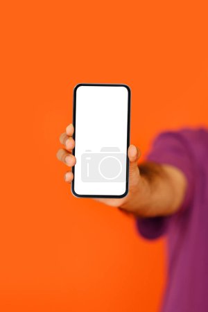 Foto de Bonita oferta en línea, aplicación móvil. Smartphone moderno con pantalla blanca en blanco maqueta en mano de hombre de piel oscura, aislado sobre fondo de estudio naranja, plano vertical recortado - Imagen libre de derechos