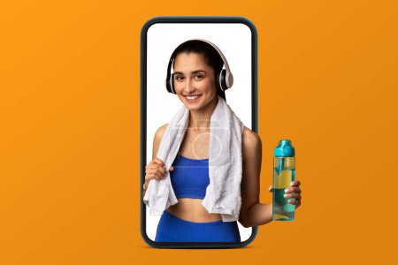 Photo for Smiling young arabic slim woman athlete in sportswear and headphones hold bottle of water on smartphone screen isolated on orange studio background. Body care, app for weight loss and fitness - Royalty Free Image