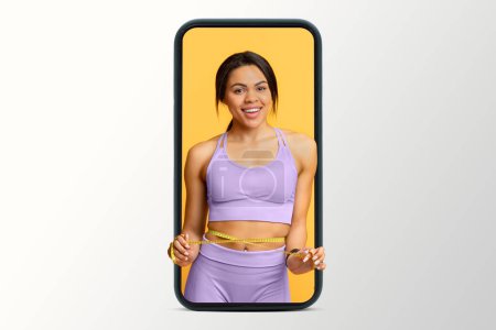 Photo for Cheerful young african american slim woman in sportswear measuring waist with measuring tape on smartphone screen isolated on white studio background. App for weight loss, body care and fitness - Royalty Free Image