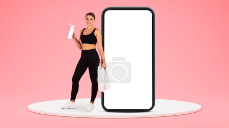Photo for Cheerful young european woman in sportswear with towel resting with water bottle on platform near big smartphone with empty screen isolated on pink studio background. App for sports, fitness - Royalty Free Image