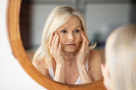 Gealterte Hautprobleme. Aufgebrachte Seniorin sah in den Spiegel und berührte Gesicht, bemerkte Falten in der Nähe der Augen, unglücklich über schlechte Hautpflege Routine und Behandlung