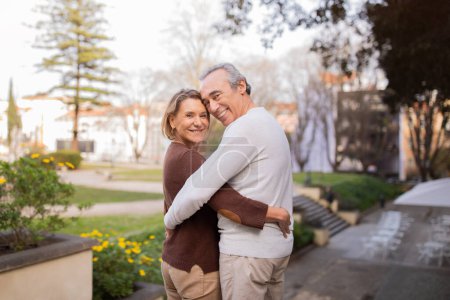 Photo for Happy Retirement Life. Mature Spouses Hugging Standing In Park In Lisbon City, Smiling To Camera. Senior Couple In Love Sharing Embrace Expressing Their Happiness, Enjoying Weekend Outside - Royalty Free Image