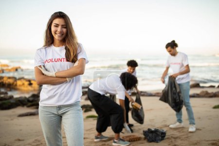 Téléchargez les photos : Heureux jeunes diverses personnes bénévoles en t-shirts et dame asiatique avec des sacs poubelles nettoyer les ordures sur la plage, en plein air. Pollution des océans et conservation de l'environnement, protection de la planète, Jour de la Terre - en image libre de droit