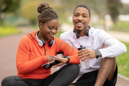 Photo for Positive young african american lady and guy checking pulse on fit tracker, enjoy workout result in park outdoor. Water for body care, cardio workout in city, fitness together with device - Royalty Free Image