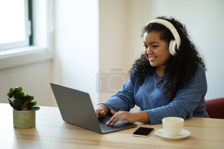 Cheerful pretty hispanic young brunette curly woman in casual sitting at desk next to window, using wireless headphones and laptop at home, typing on computer keyboard, drinking coffee, copy space