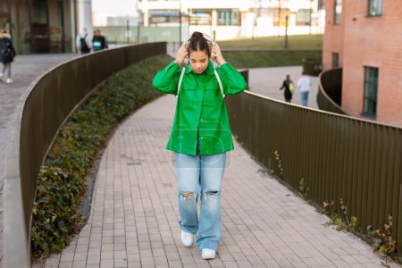 Photo for Teen student girl walking near university building outdoors, wearing headphones, spending free time after classes outside. College lady walking at campus - Royalty Free Image
