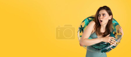 Foto de Mujer joven sorprendida en traje de baño sosteniendo anillo inflable y mirando a un lado en el espacio libre con emoción, posando sobre fondo amarillo vivo, panorama. Concepto de verano, hotel y descanso marino - Imagen libre de derechos
