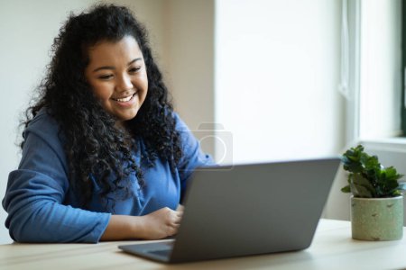 Photo for Freelance, remote job concept. Happy young chubby hispanic lady entrepreneur sitting at table, working from home, using laptop, looking at computer screen and smiling, copy space - Royalty Free Image
