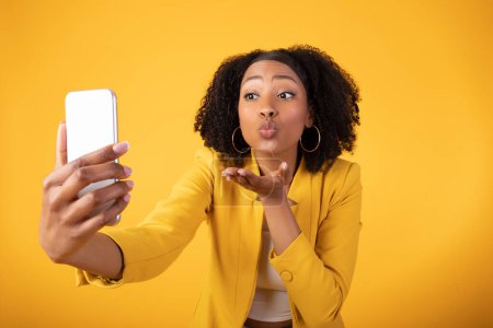 Foto de Flirty dama negra tomando selfie con el teléfono celular, el envío de beso de aire y la filmación de contenido para el blog, posando en el fondo del estudio amarillo. Mujer joven charlando en línea con el novio - Imagen libre de derechos