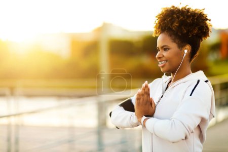 Photo for Fresh-Air Yoga. Portrait Of Young African American Woman Meditating Outdoors, Smiling Sporty African American Female In Earphones Exercising Outside, Holding Hands In Namaste Gesture, Sun Flare - Royalty Free Image