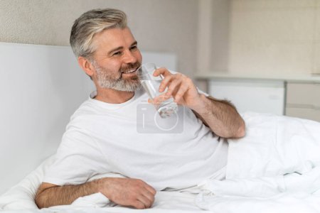 Téléchargez les photos : Positif joyeux beau mature aux cheveux gris homme européen portant un pyjama assis au lit, tenant un verre d'eau, buvant de l'aqua après le réveil le matin et souriant, intérieur de la maison, espace de copie - en image libre de droit