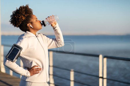 Téléchargez les photos : Fatiguée sportive noire athlète féminin buvant de l'eau de la bouteille après l'entraînement en plein air, assoiffée athlétique afro-américaine debout sur la jetée près de la mer, prendre une pause pendant l'entraînement de remise en forme à l'extérieur - en image libre de droit