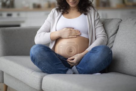 Foto de Edad Materna Avanzada. Mujer embarazada sonriente sentada en el sofá en casa y abrazando el vientre, tiro recortado de embarazada acariciando tiernamente al bebé, disfrutando del tiempo de espera, primer plano - Imagen libre de derechos