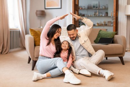 Téléchargez les photos : Real Estate Dream. Famille asiatique joyeuse avec petite fille joignant les mains au-dessus de bébé faisant toit symbolique, posant assis sur le sol ensemble à la maison. Acheteurs de maisons Planification Achat de maisons - en image libre de droit
