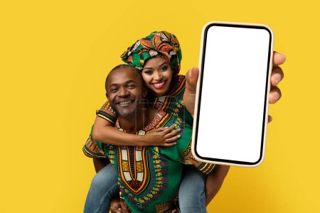 Photo for Cheerful pretty young black woman piggybacking her happy middle aged boyfriend, showing cell phone with white blank screen, couple wearing african costumes, mockup on yellow background - Royalty Free Image
