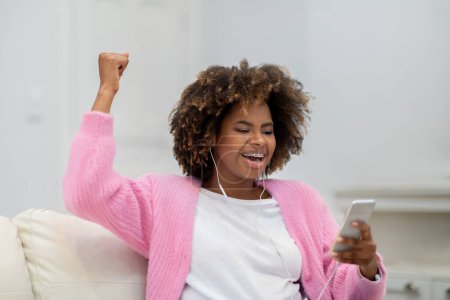 Téléchargez les photos : Enthousiaste femme noire bouclée émotionnelle millénaire avec des appareils dentaires assis sur le canapé dans le salon confortable, en utilisant smartphone et écouteurs, regarder le jeu en ligne, geste, espace de copie - en image libre de droit