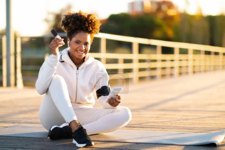 Photo for Fitness Snacks. Smiling Black Woman Eating Proteing Bar After Training Outdoors, Happy Young African American Female Athlete Enjoying Healthy Snack And Using Smartphone While Relaxing Outside - Royalty Free Image