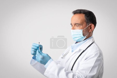 Photo for Serious european senior doctor in white coat, protective mask and gloves with syringe on gray background. Vaccination, treatment of disease and injection, medical care, ad and offer - Royalty Free Image