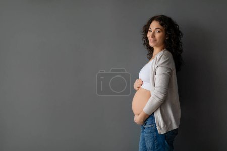 Foto de Perfil Shot of Beautiful Pregnant Woman abrazando tiernamente el vientre mientras posaba contra la pared del estudio gris, feliz madre embarazada joven acariciando el golpe del bebé y sonriendo a la cámara, espacio de copia - Imagen libre de derechos
