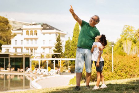 Téléchargez les photos : Souriant vieux caucasien grand-père étreindre petit garçon métis pointant du doigt à l'espace vide, profiter du temps libre, s'amuser en plein air. Repos, week-end et vacances dans le parc ensemble, relation familiale - en image libre de droit
