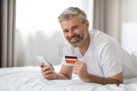 Photo for E-commerce, retail. Cheerful handsome middle aged grey-haired man with beard lying on bed, using smartphone and plastic bank card, making online order while staying home at weekend, copy space - Royalty Free Image