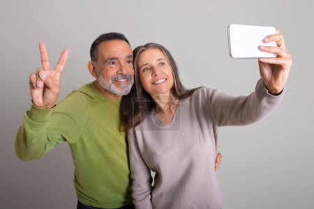 Téléchargez les photos : Homme âgé caucasien souriant et dame pointant signe de paix avec les doigts et prendre selfie sur smartphone sur fond de studio gris. Relation et amour, photo pour réseau social et blog - en image libre de droit