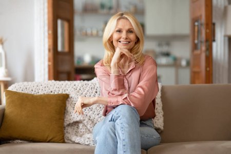 Happy Blonde Mature Woman Enjoying Her Time At Home, Posing Sitting On A Cozy Couch And Smiling To Camera Resting Chin On Hand. Portrait Of Confident Lady. Domestic Comfort Concept