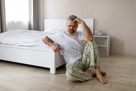 Photo for Upset pensive middle aged grey-haired handsome man wearing pajamas sitting on floor by bed at home, touching head, suffering from hangover or anxiety, copy space. Permacrisis, midlife crisis - Royalty Free Image