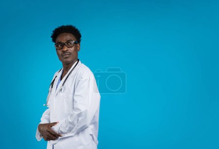 Téléchargez les photos : Beau jeune médecin afro-américain confiant portant des lunettes, un manteau médical et avec un stéthoscope sur le cou posant sur fond bleu, regardant l'espace de copie. Concept de soins de santé - en image libre de droit