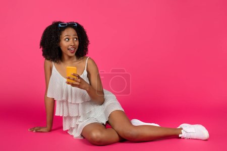 Téléchargez les photos : Souriant jeune femme frisée afro-américaine mignonne en robe bavarder sur smartphone, assis sur le sol, regardant l'espace vide, isolé sur fond de studio rose. Réseaux sociaux, surprise, annonce et offre - en image libre de droit