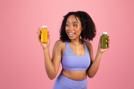 Foto de Sorprendido feliz millennial mujer afroamericana en ropa deportiva sostienen frascos de jugo o batidos aislados sobre fondo rosa, estudio. Cuidado corporal, vitaminas para el deporte, dieta y oferta - Imagen libre de derechos