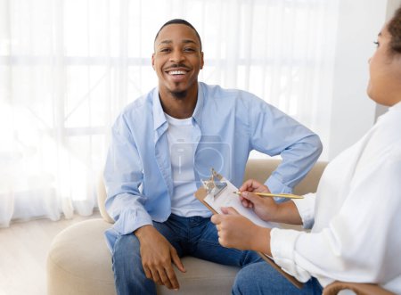 Photo for Positive happy young african american guy in casual sitting on couch against woman psychologist, male patient smiling at camera during session with therapist, successful therapy concept - Royalty Free Image