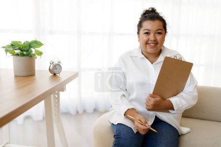 Foto de Feliz sonriente hermosa joven mestiza dama de raza más tamaño vistiendo traje formal psicólogo posando en la clínica acogedora oficina, sentado en el sofá, sosteniendo la carpeta en su mano, espacio de copia - Imagen libre de derechos